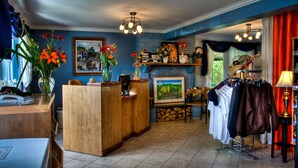 A reception area with a wooden desk, a phone, and a vase with flowers. There is a painting on the wall and a shelf with various items. - Motel Le JoliBourg (Saint-Sauveur)