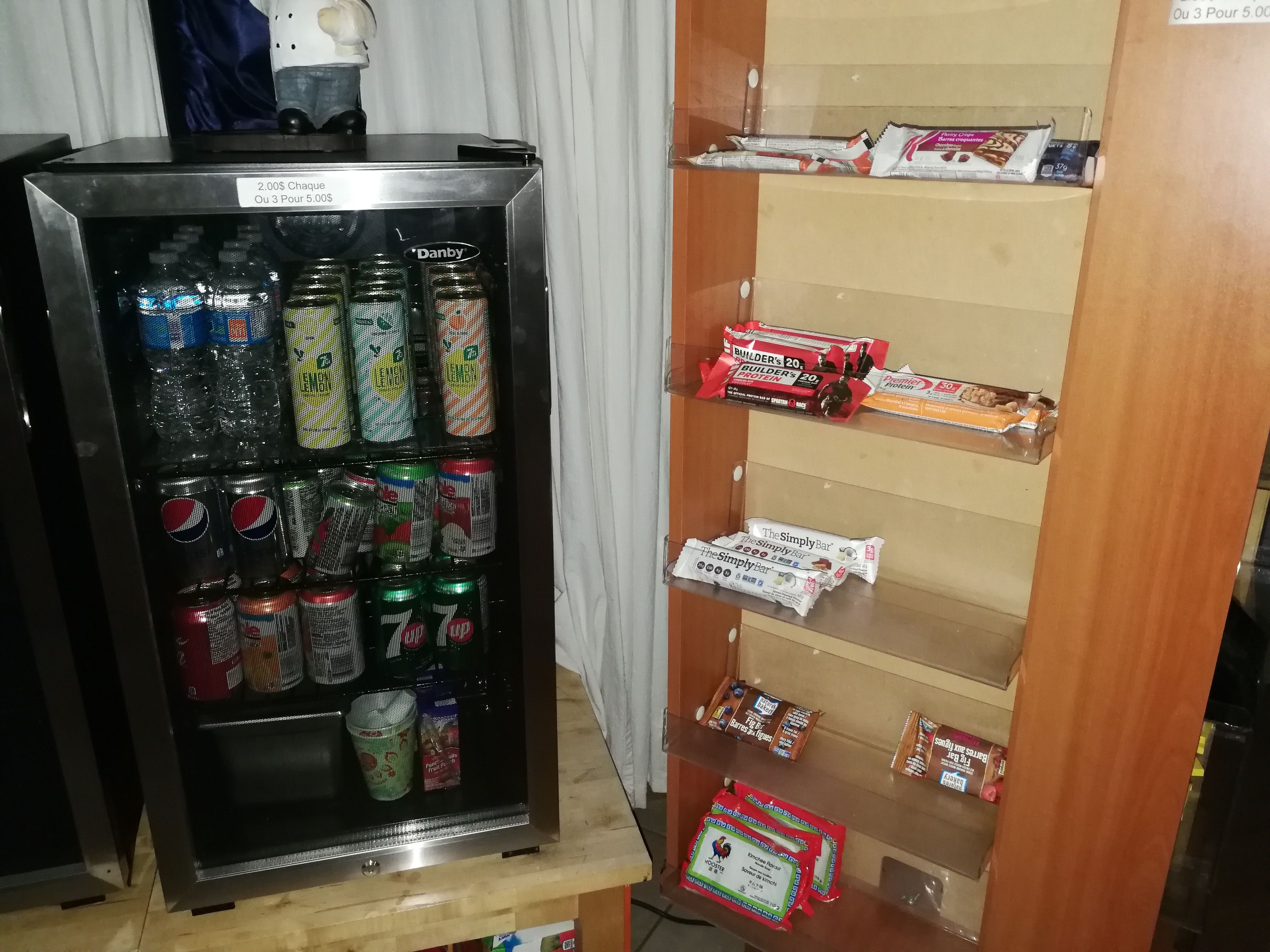 Máquina expendedora repleta de diversas bebidas y aperitivos, incluyendo latas y botellas, y un estante con alimentos envasados.