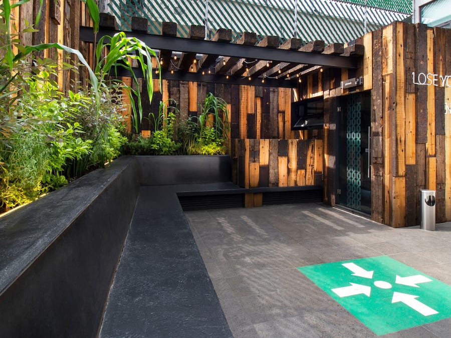 Entrada de un edificio moderno con fachada de madera, una flecha direccional verde en el suelo y una frase inspiradora en la pared.