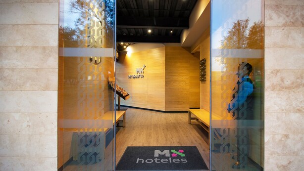 A hotel entrance with a wooden reception desk, a bench, and a doormat with the hotel's logo. - Hotel MX aeropuerto CDMX, Trademark Collection by Wyndham (Mexico City)
