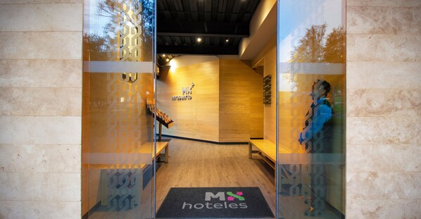 Entrada de un hotel con un mostrador de recepción de madera, un banco y un felpudo con el logotipo del hotel.