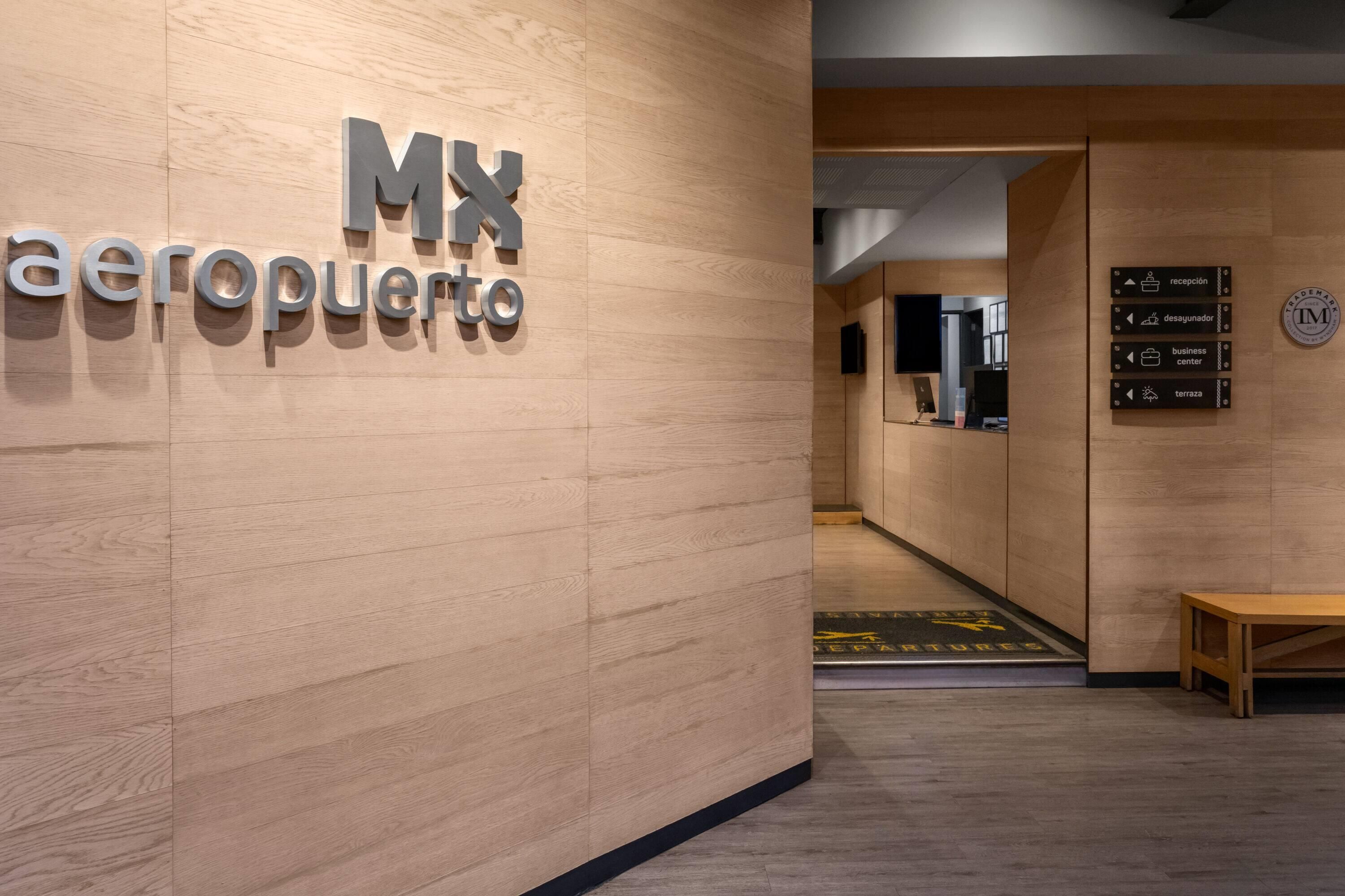 Un interior de madera con una mesa de recepción, un cartel en la pared que dice 'MX aeroporto' y señales direccionales para 'recepción', 'desayunador', 'centro de negocios' y 'terrazas'.