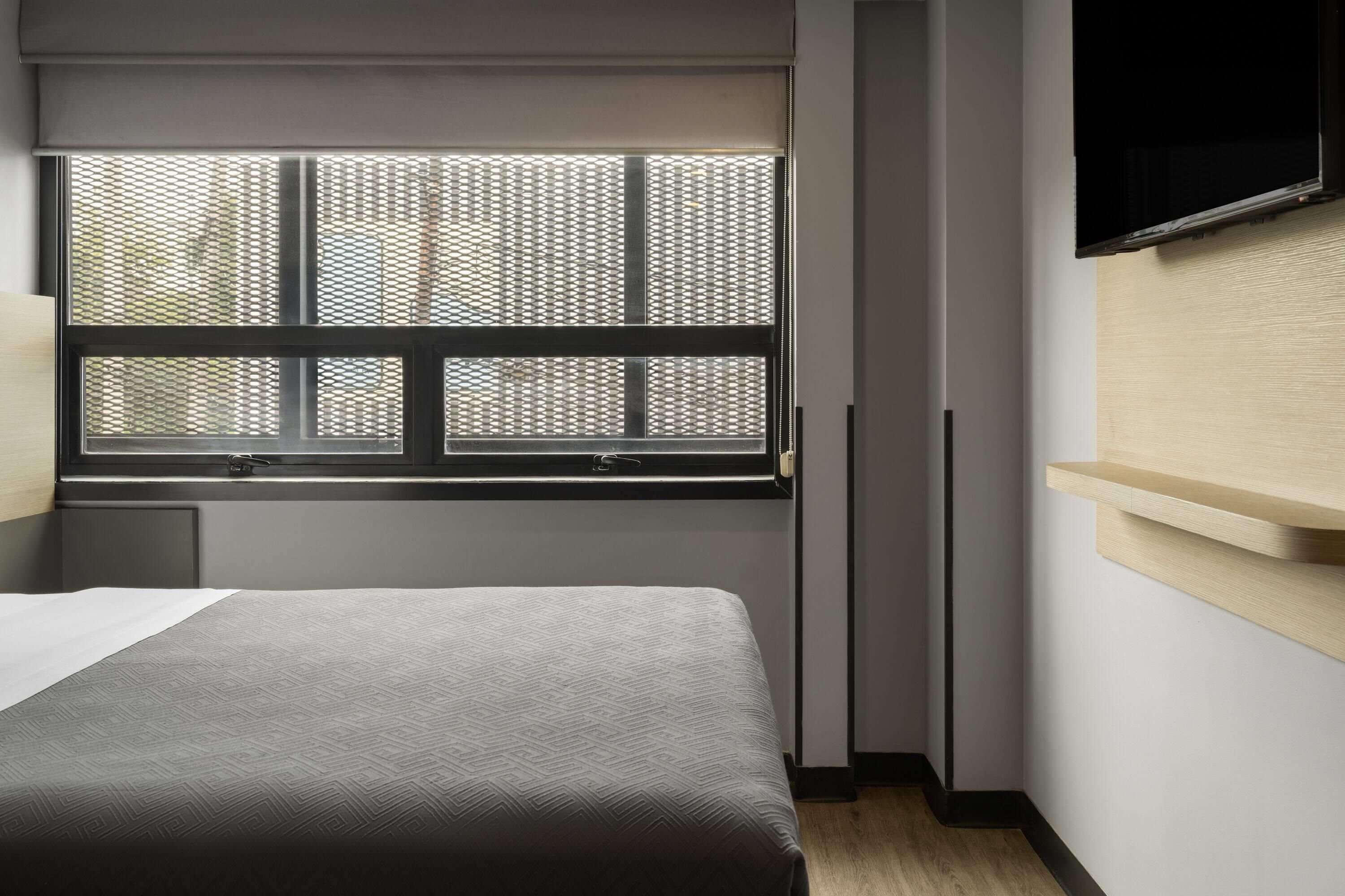 Quarto com cama, janela com persianas, televisão fixada na parede e piso de madeira.