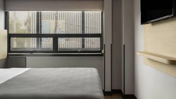 Quarto com cama, janela com persianas, televisão fixada na parede e piso de madeira.
