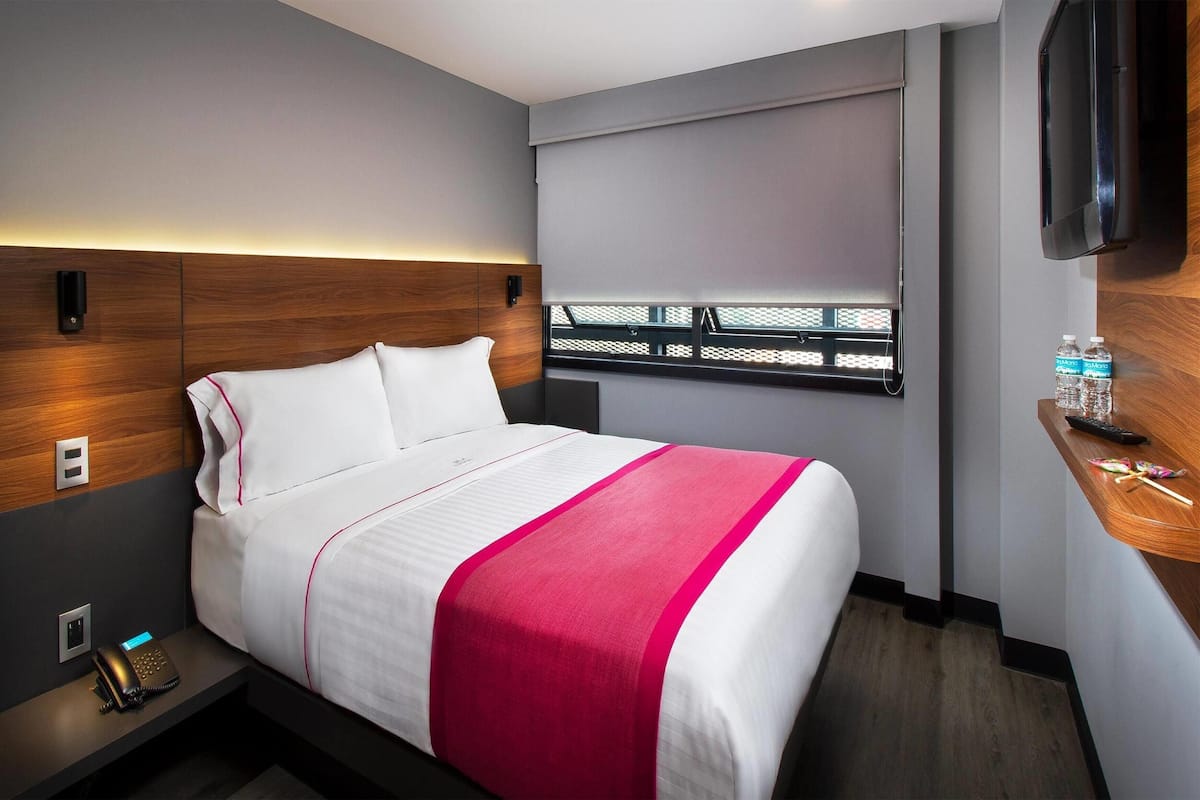 A modern hotel room with a neatly made bed, a bedside table with a phone, a TV mounted on the wall, and a window with a view.