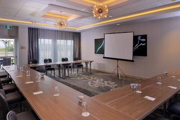 Une salle de conférence avec une longue table, des chaises, un écran de projection et une grande fenêtre avec des rideaux.