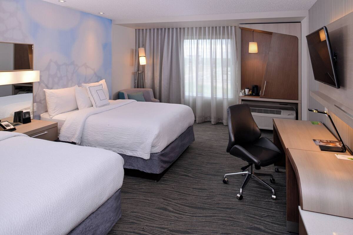 A hotel room with two beds, a desk with a chair, a flat-screen TV, a desk lamp, a coffee maker, and a large window with curtains.