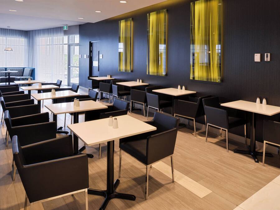 Een moderne eetruimte met meerdere tafels en stoelen, grote ramen met gele gordijnen, en een strak, eigentijds design.