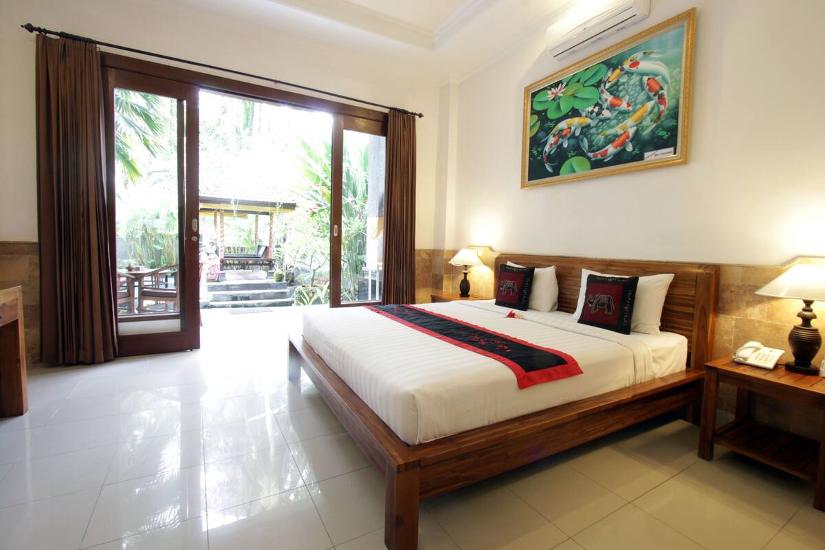 A hotel room with a large bed, wooden headboard, two bedside tables with lamps, a painting on the wall, and a view of the outdoors through a sliding door.