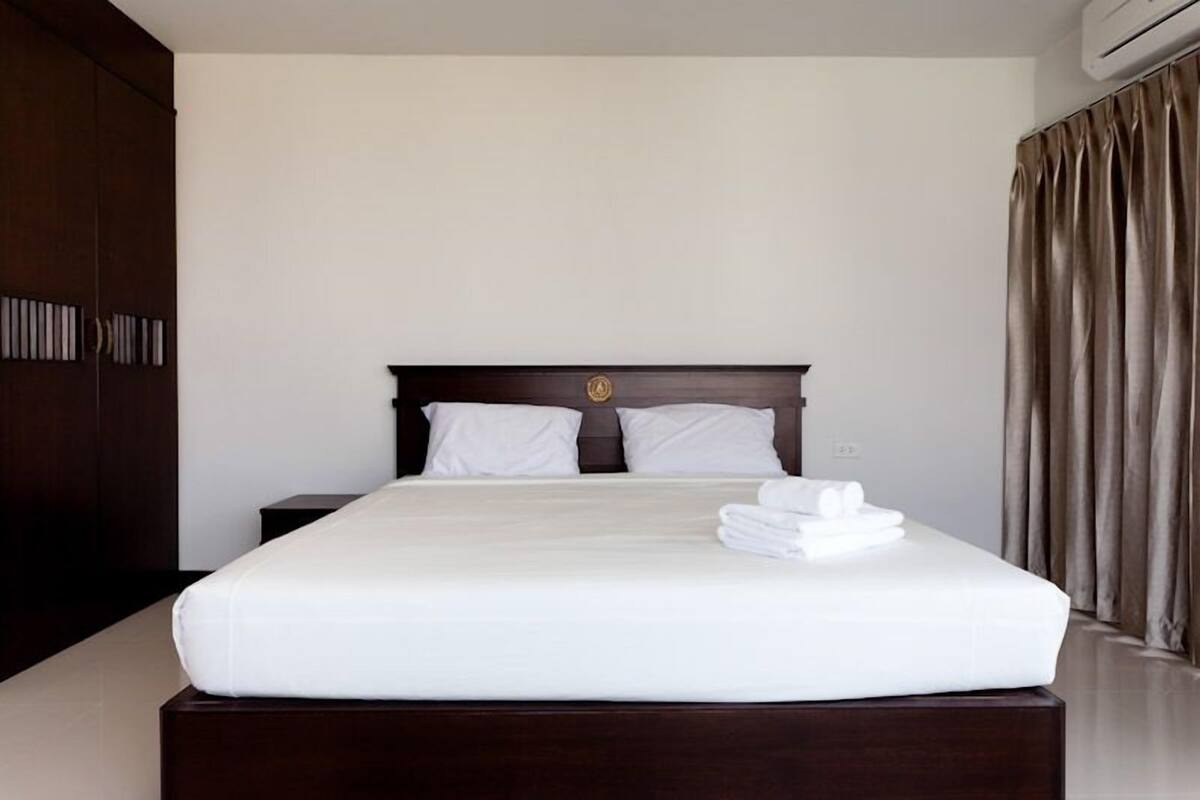 A neatly made bed with white linens and a folded towel, a dark wooden headboard, and a large window with curtains.