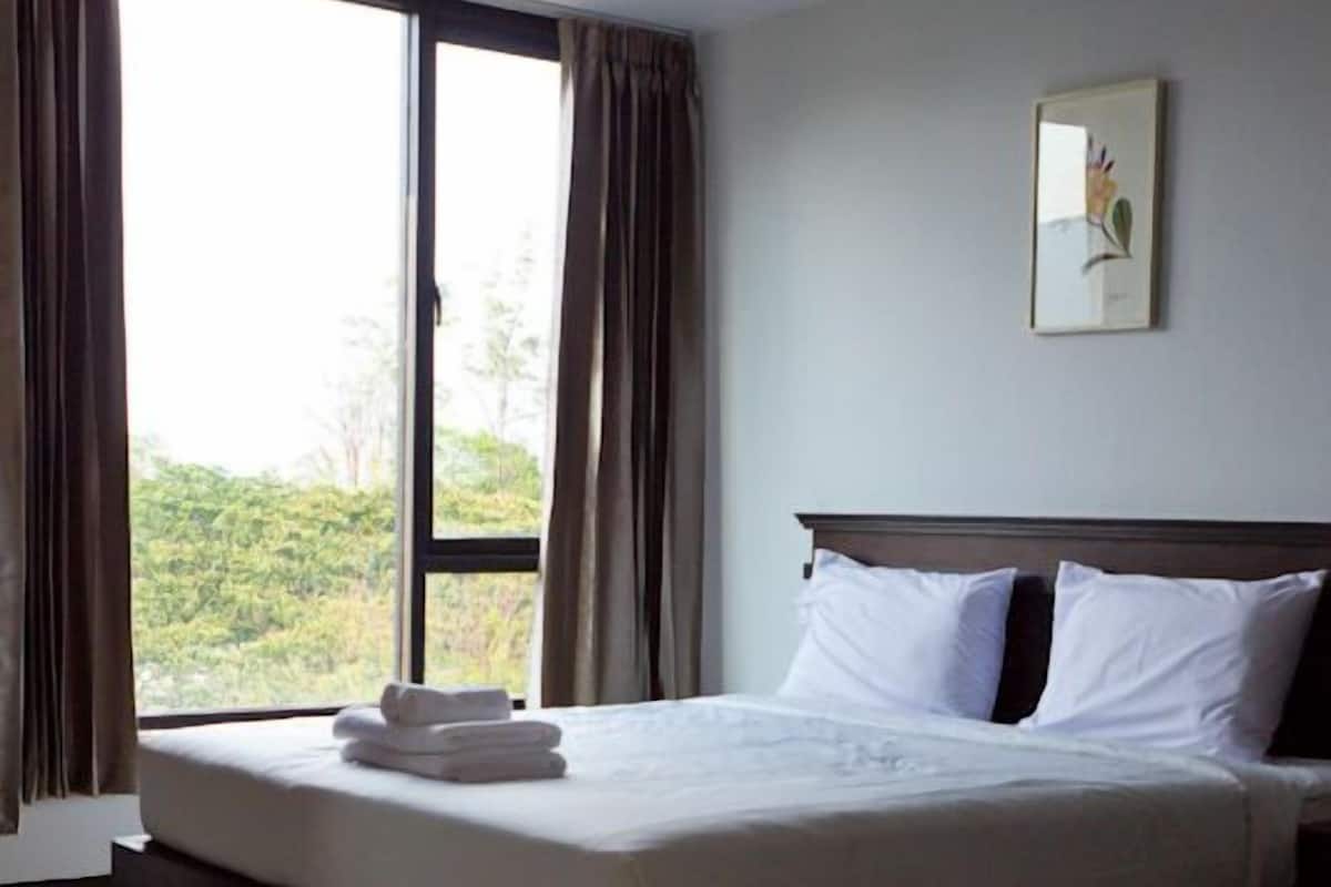 A neatly made bed with white linens and a folded towel, a large window with curtains, and a framed picture on the wall.
