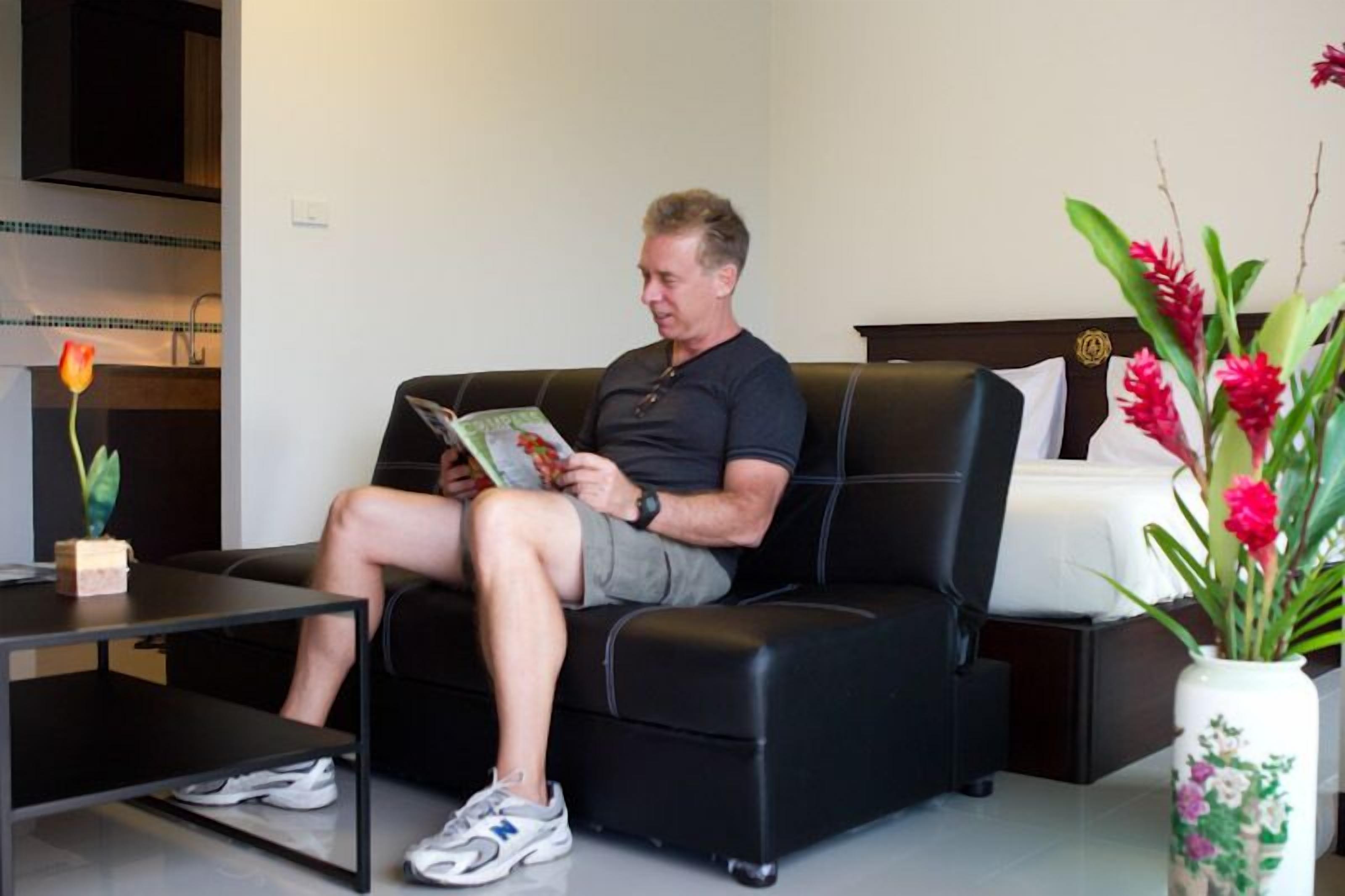 Ein Mann sitzt auf einer schwarzen Couch in einem modernen Zimmer mit einem weißen Bett, einem kleinen Tisch und einer Vase mit Blumen.