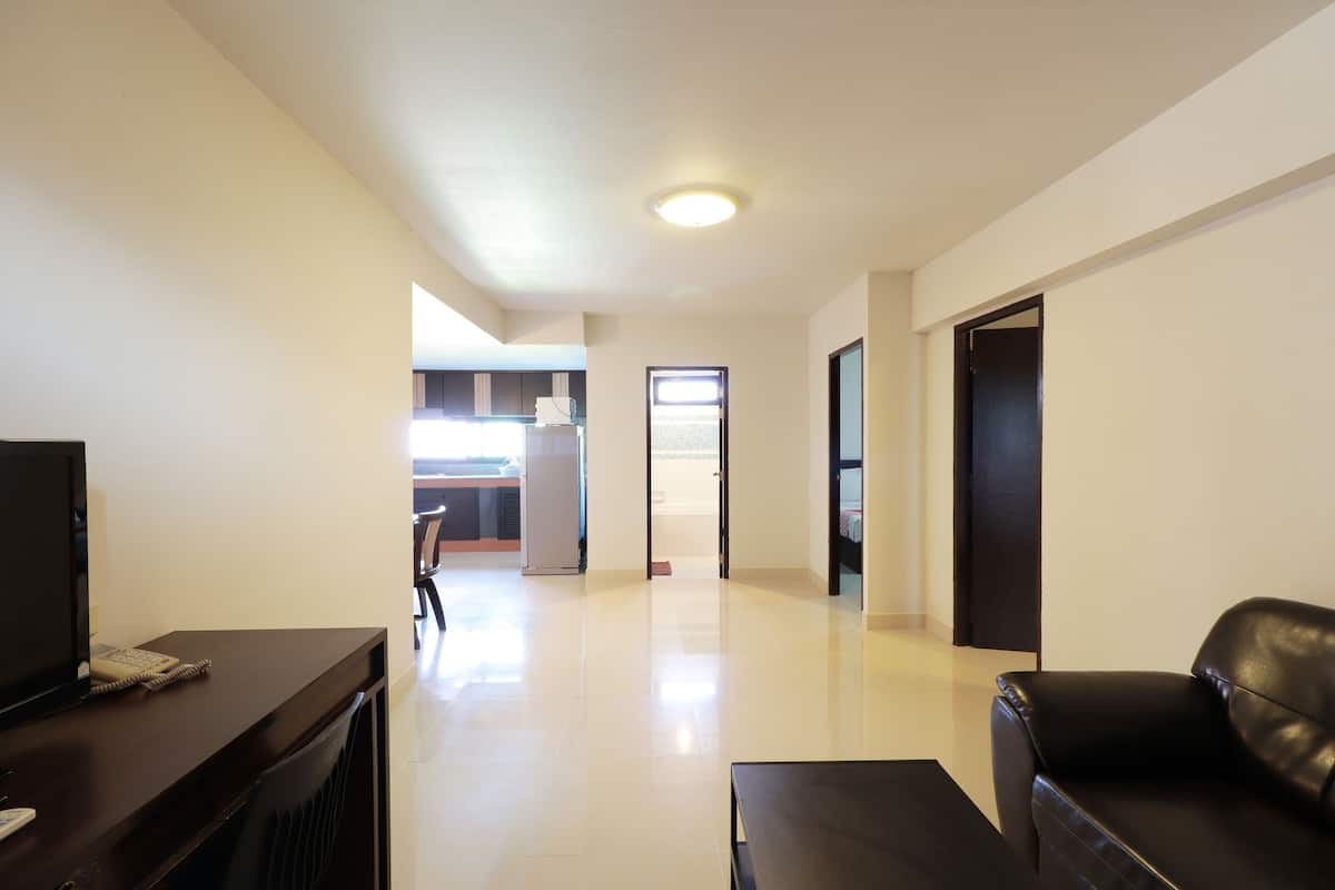 A spacious living area with a flat-screen TV, a dark wooden coffee table, and a black leather sofa. A kitchen area is visible in the background with a refrigerator and a window.