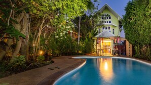 Una piscina de borde curvo, rodeada de abundante vegetación y un edificio de estilo arquitectónico tradicional.