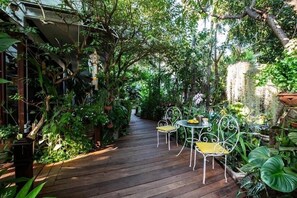 Un sendero de madera rodeado de abundante vegetación que lleva a un pequeño espacio al aire libre con sillas y una mesa de metal.