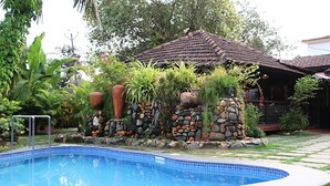 Una piscina con borde de piedra y plantas, una estructura de techo con tejas y un camino de piedra.
