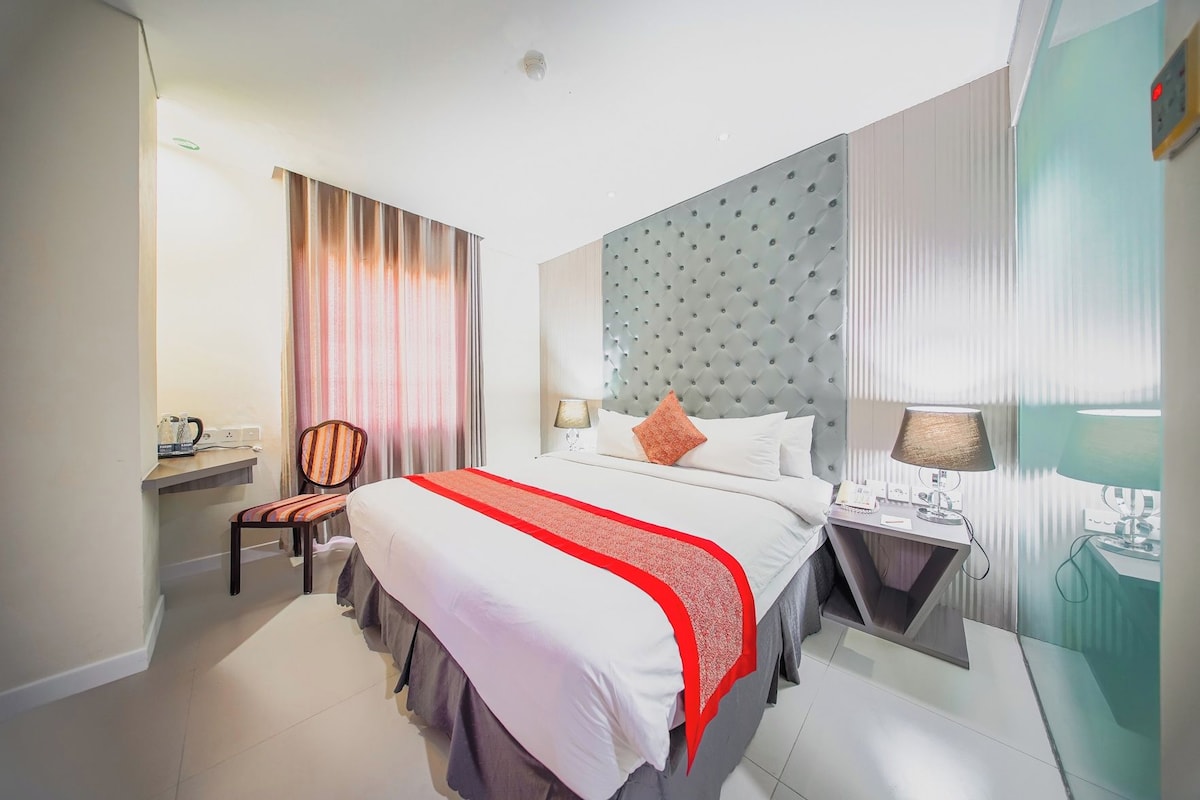 A hotel room with a bed, a chair, a bedside table, a lamp, and a mirror.