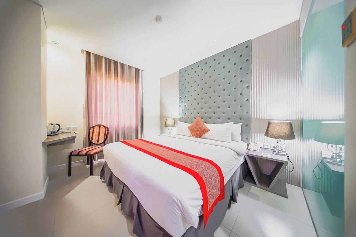 Ein Hotelzimmer mit einem großen Bett, einem Nachttisch mit Lampe, einem Sessel, einem Schreibtisch mit Computer und einem Fenster mit Vorhängen.