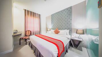 Ein Hotelzimmer mit einem großen Bett, einem Nachttisch mit Lampe, einem Sessel, einem Schreibtisch mit Computer und einem Fenster mit Vorhängen.