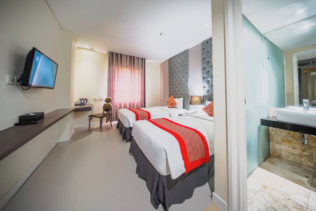 Ein Hotelzimmer mit zwei Betten, einem Fernseher, einem Schreibtisch mit Stuhl, einem Badezimmer inklusive Spiegel und Duschbereich.