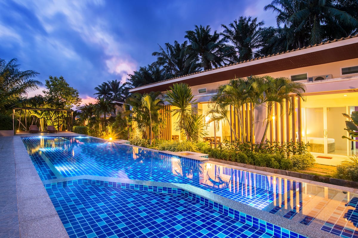 A swimming pool with a tiled edge, surrounded by palm trees and a building with a balcony.