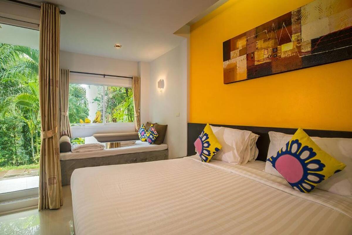 A hotel room with a large bed, a window with a view of greenery, and a yellow accent wall.