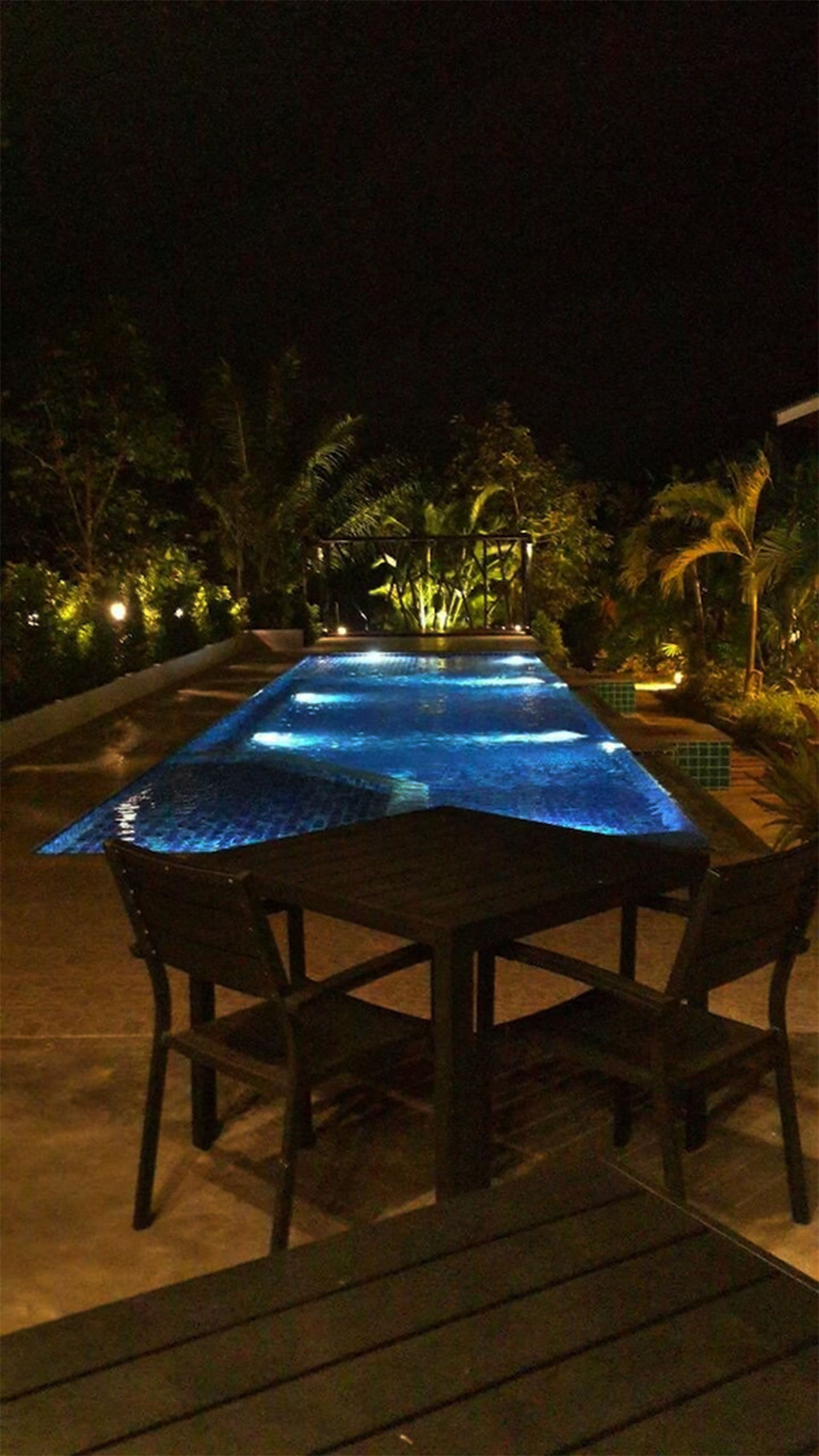 Un área de comedor al aire libre con una mesa cubierta por un mantel iluminado en azul, rodeada de sillas, con follaje y palmeras de fondo.