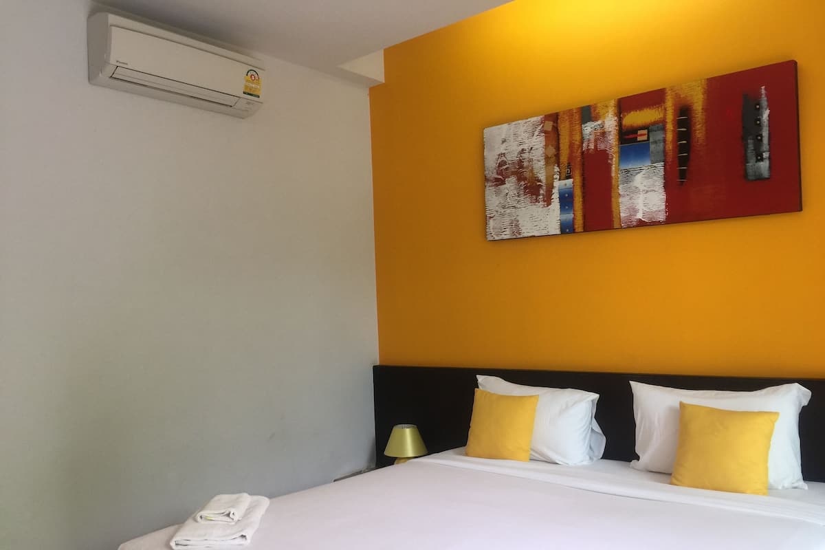 A hotel room with a bed, white bedding, a yellow pillow, a white pillow, a black headboard, and a wall-mounted air conditioner.