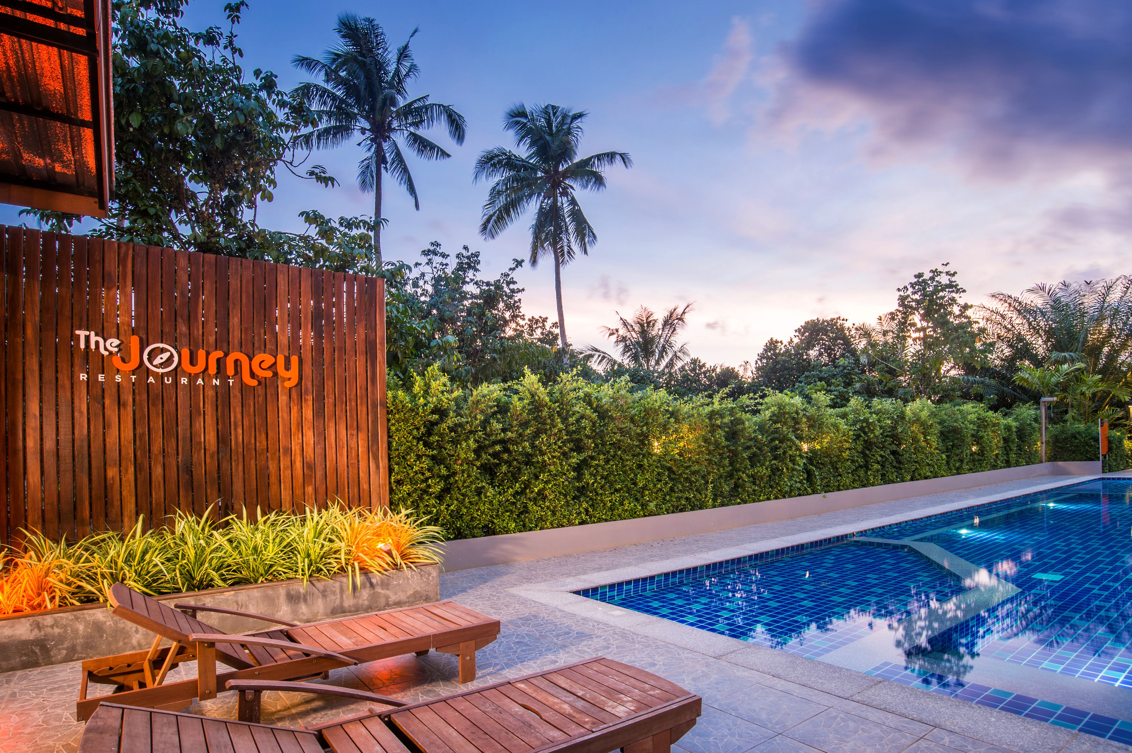 A poolside area with wooden loungers, a wooden fence, and lush greenery including palm trees.