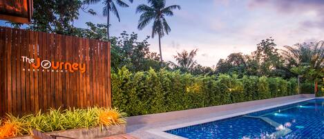A poolside area with wooden loungers, a wooden fence, and lush greenery including palm trees.