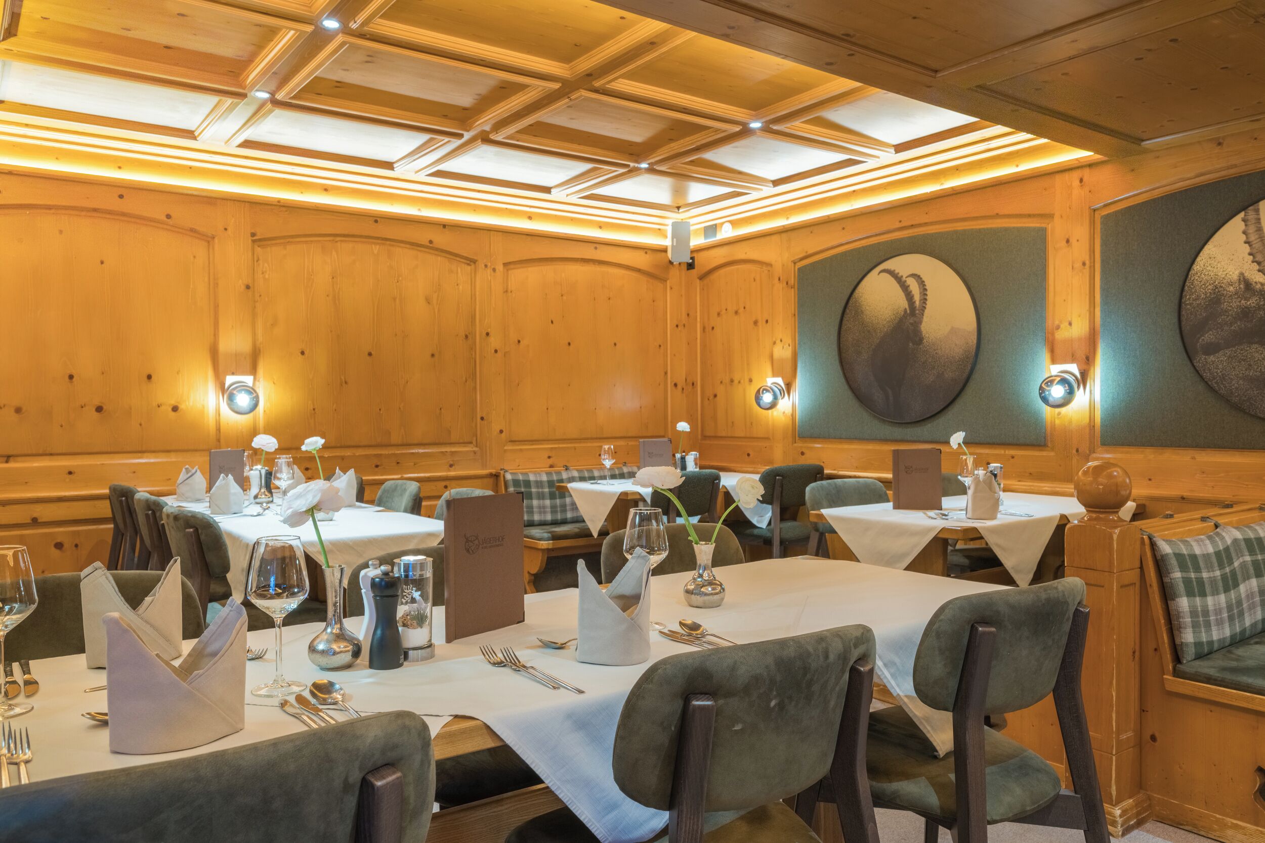 Ein Restaurant mit Holzwandverkleidung, runden Tischen mit weißen Tischdecken und grünen Polsterstühlen.