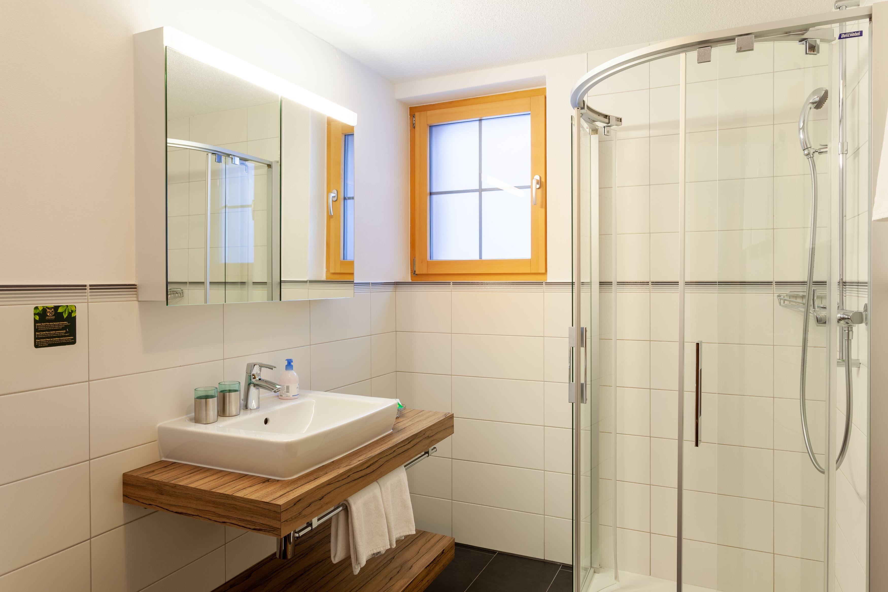 Un baño moderno con ducha envuelta en vidrio, un lavamanos blanco y un mueble de tocador de madera.