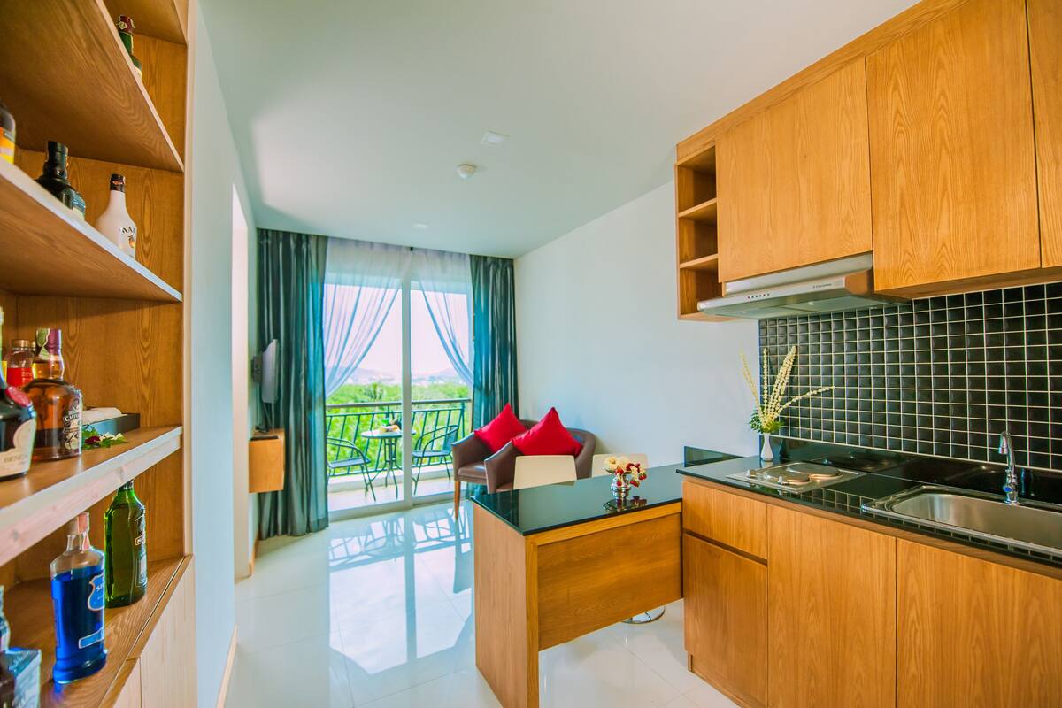 A modern kitchen with wooden cabinets, a black countertop, and a view of a balcony with green chairs.