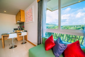 A modern kitchen with a dining area, a green sofa with colorful cushions, and a balcony with a scenic view.