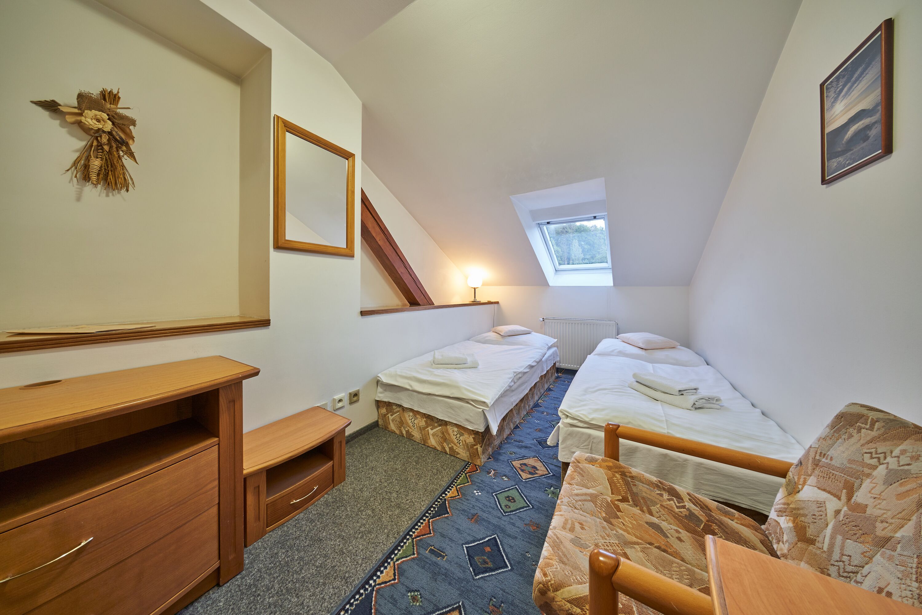 A small, well-lit room with two single beds, a wooden desk, a chair, a small table, and a window providing natural light.