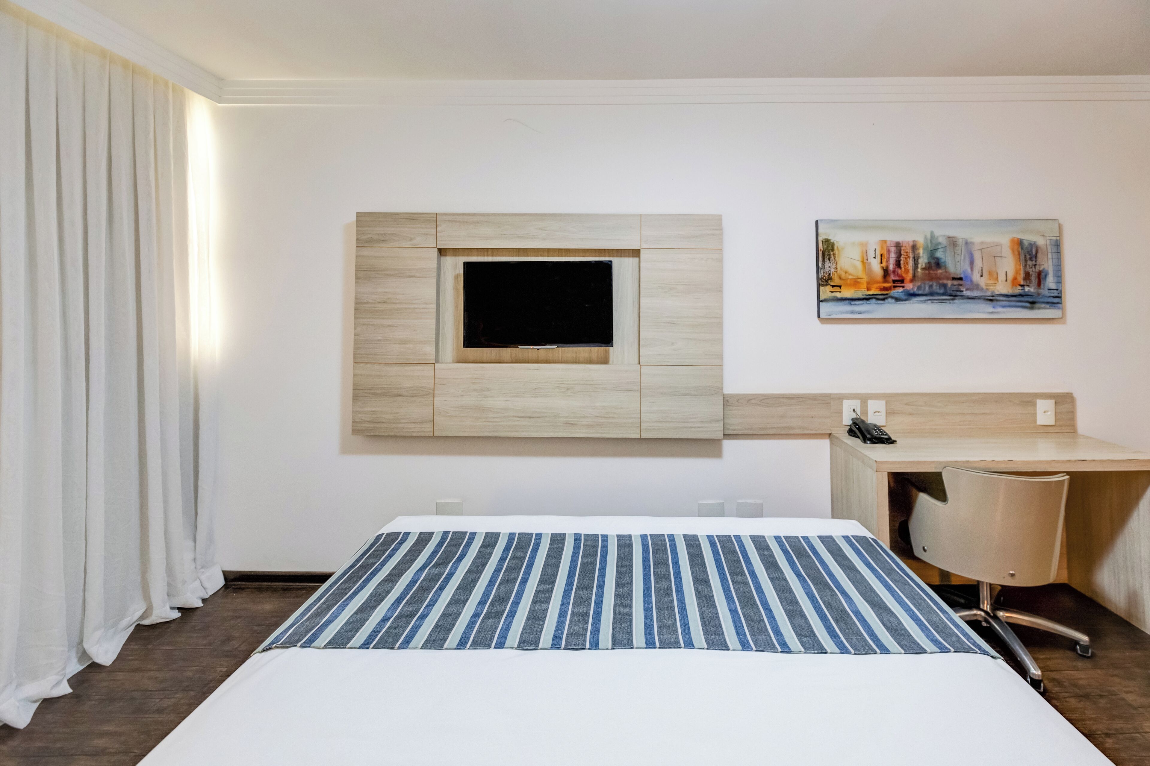 Habitación de hotel con cama, escritorio, televisor montado y un cuadro en la pared.