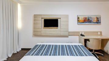 Habitación de hotel con cama, escritorio, televisor montado y un cuadro en la pared.