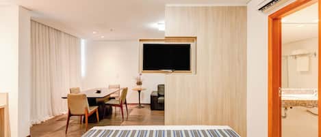A hotel room with a bed, a TV mounted on the wall, a dining area with chairs and a table, and a bathroom visible through an open door.