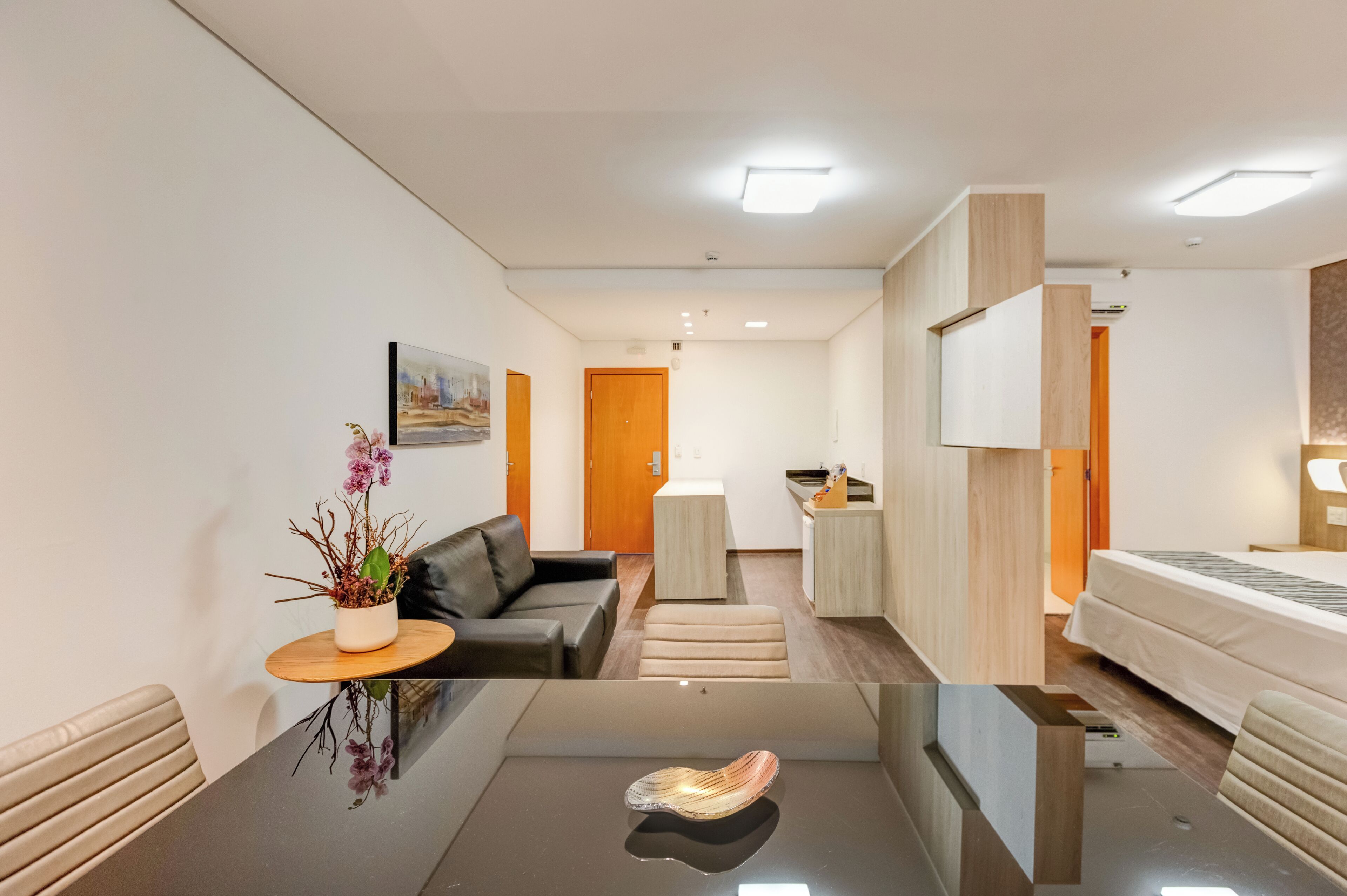 Quarto de hotel moderno com mesa de vidro, sofá, cama e uma pequena cozinha.
