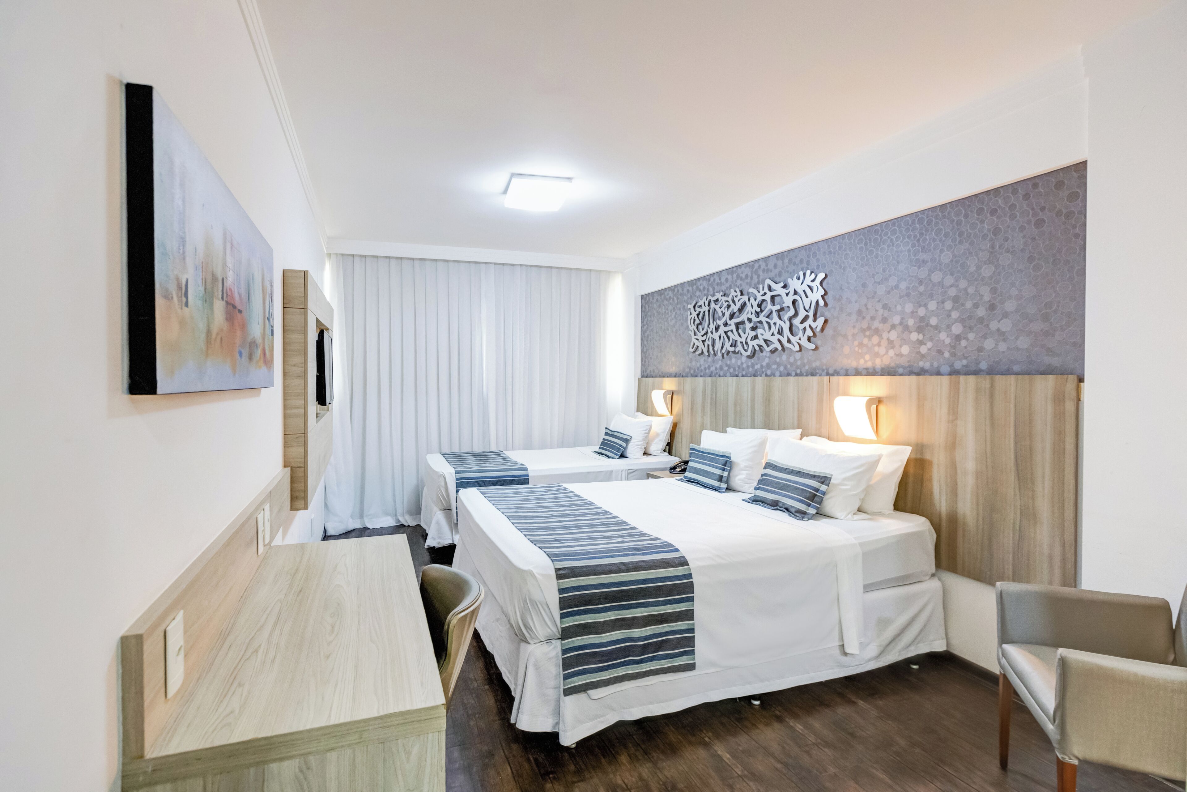 Ein Hotelzimmer mit zwei Betten, einem Schreibtisch und einem Stuhl. An der Wand hängt ein Gemälde und über den Betten ein dekoratives Wandelement.