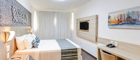 A hotel room with a bed, bedside table, desk, chair, TV, artwork, and a window with blinds.