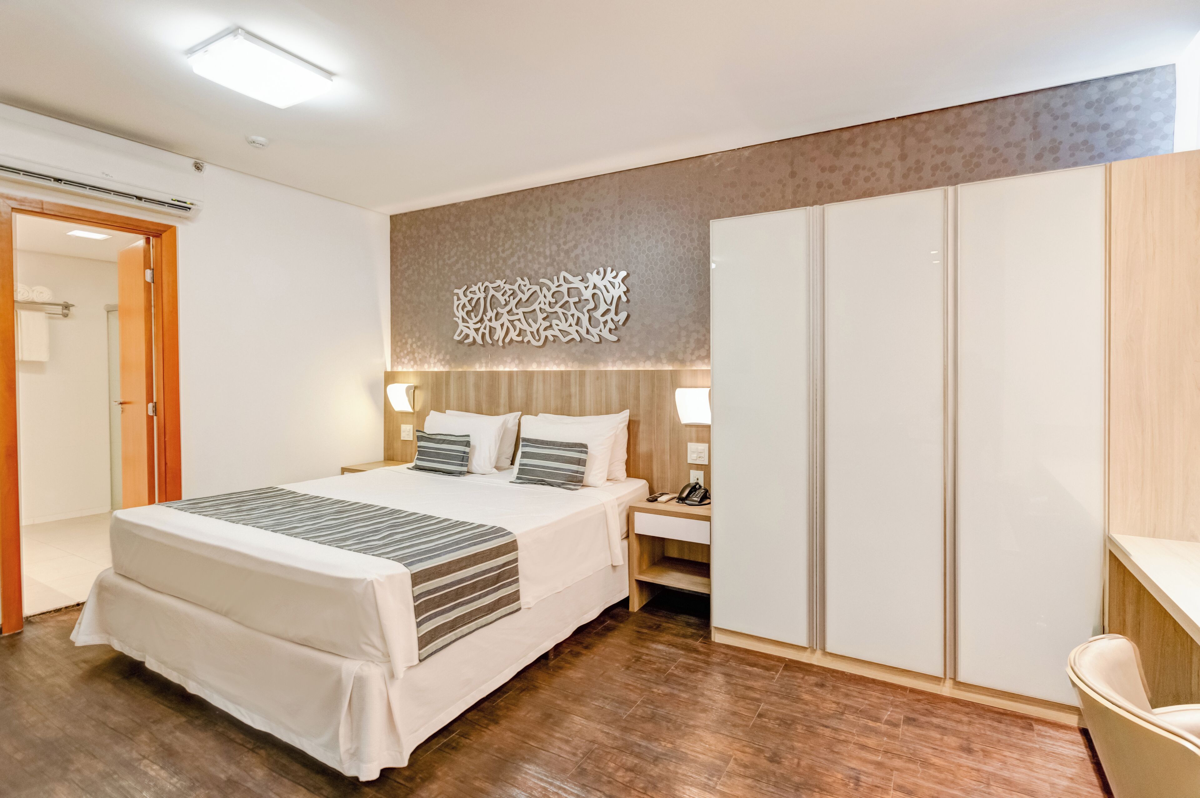A hotel room with a bed, bedside table, lamp, wardrobe, and a decorative wall piece.
