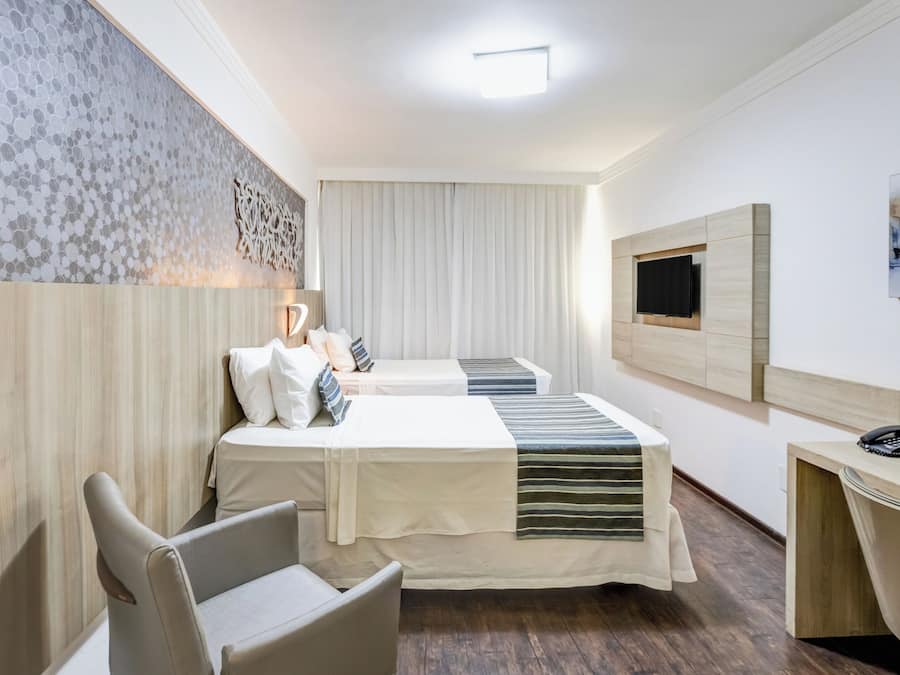 A hotel room with two beds, a desk, a chair, a TV, and a large window with curtains.
