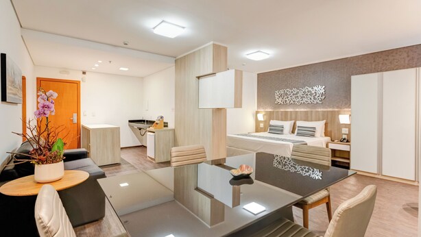 Een moderne hotelkamer met een glazen eettafel, comfortabele zitplaatsen, een bed met hoofdeinde en een decoratief wandkunstwerk.