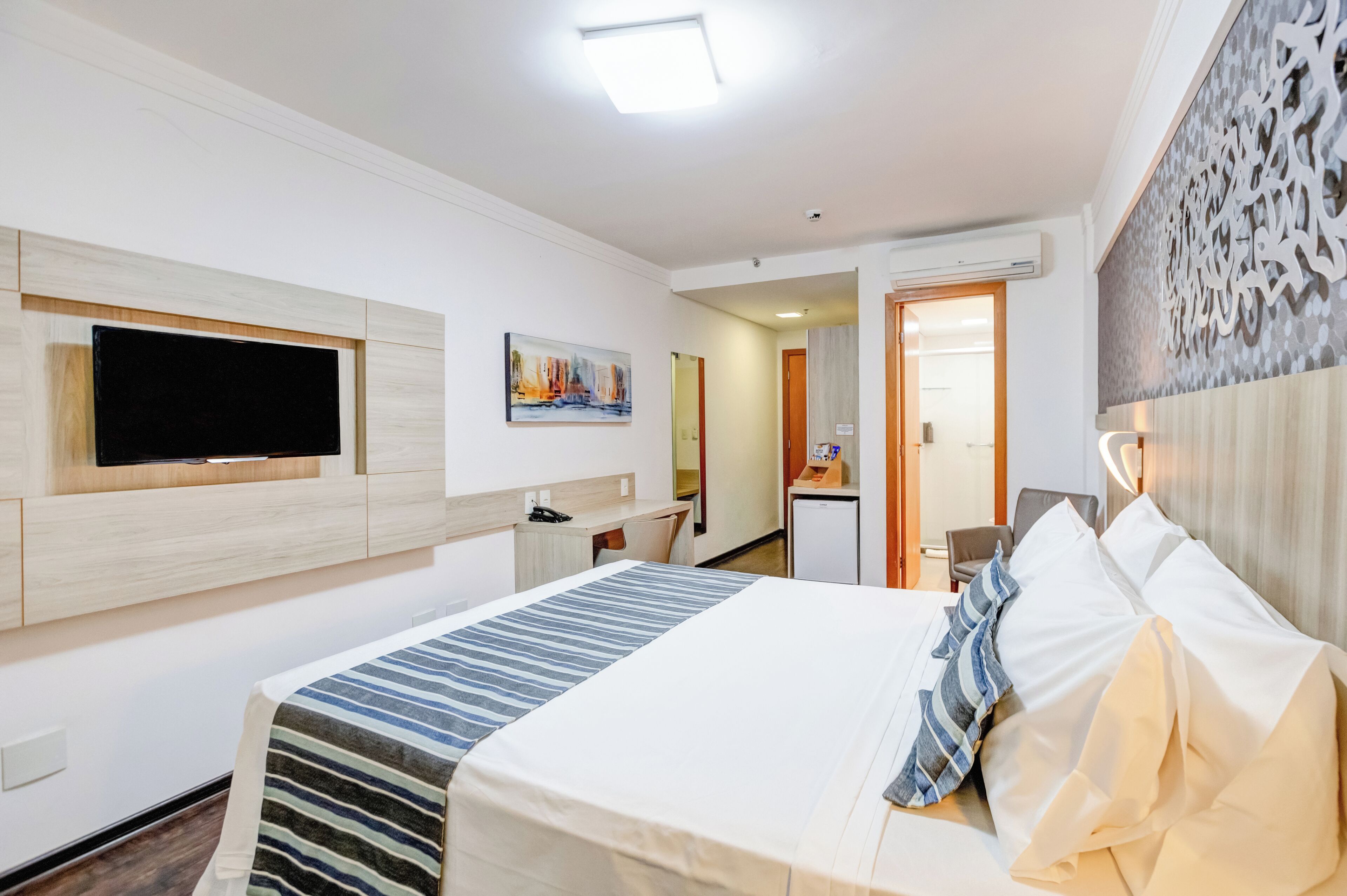 A hotel room with a bed, a flat-screen TV mounted on the wall, a desk with a chair, and a small kitchenette.