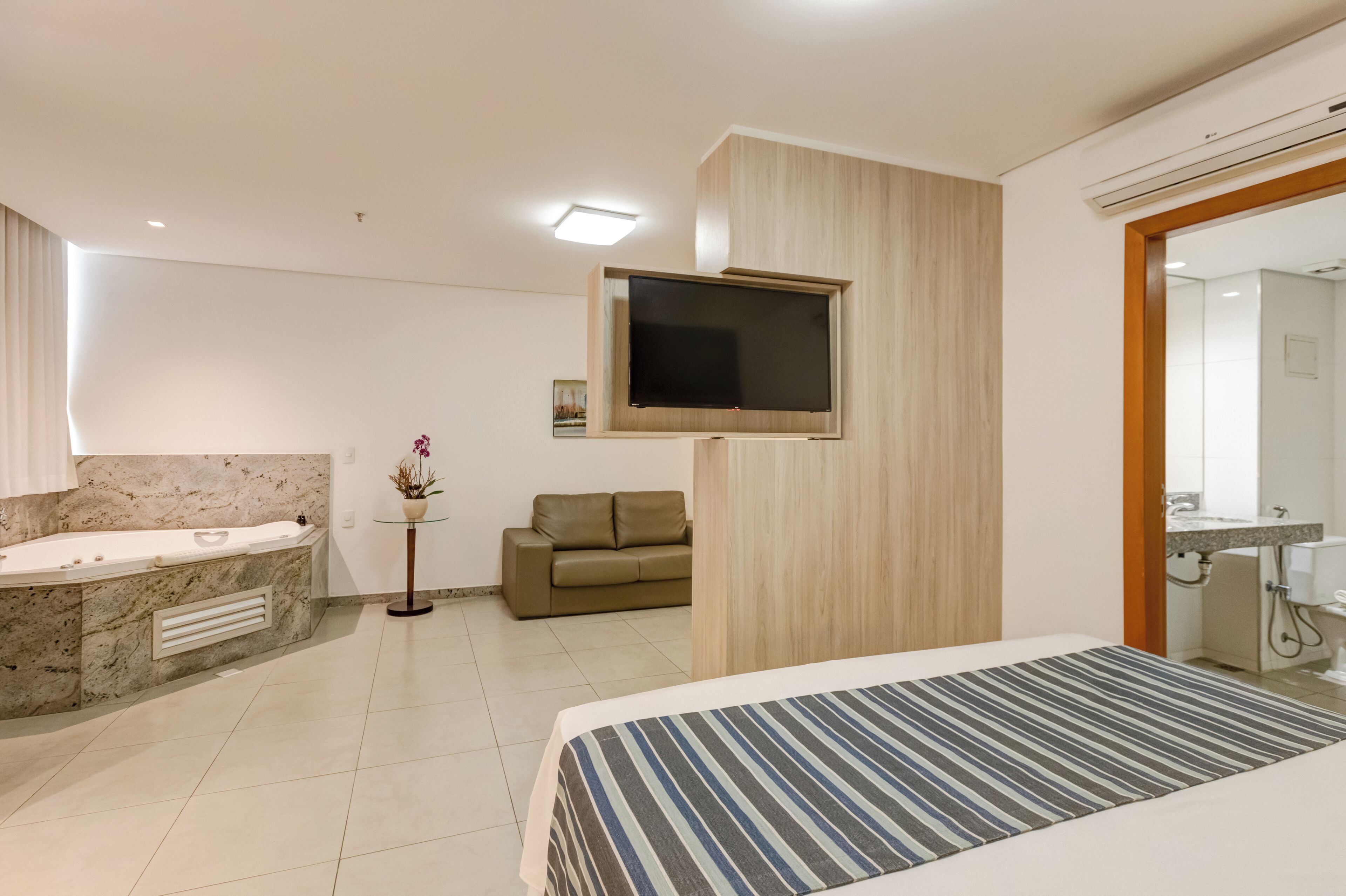 A modern hotel room with a large bed, a built-in bathtub, a sofa, a TV mounted on the wall, and a bathroom with a sink and mirror.