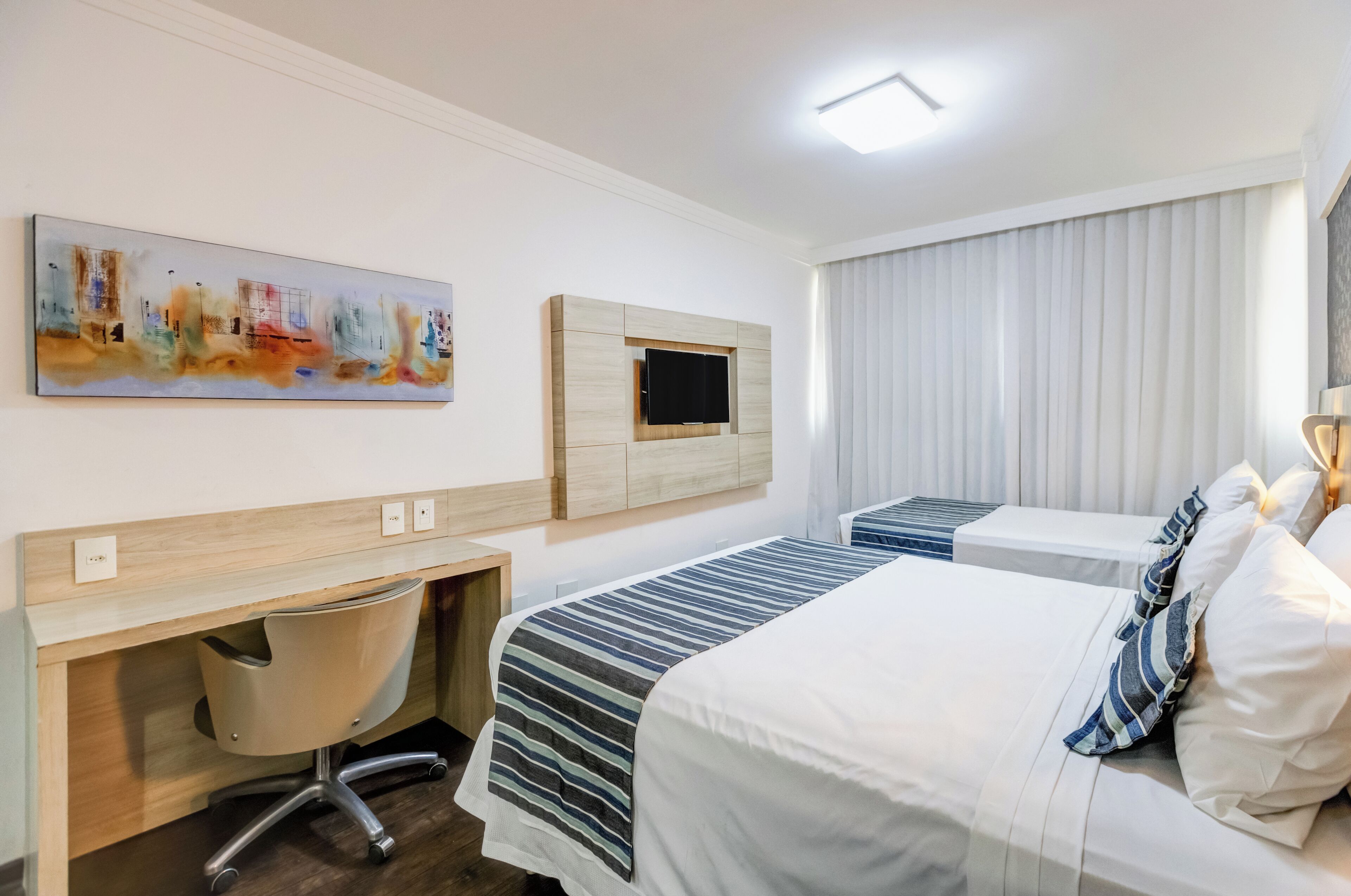 A hotel room with a bed, a desk with a chair, a TV mounted on the wall, and a window with blinds.
