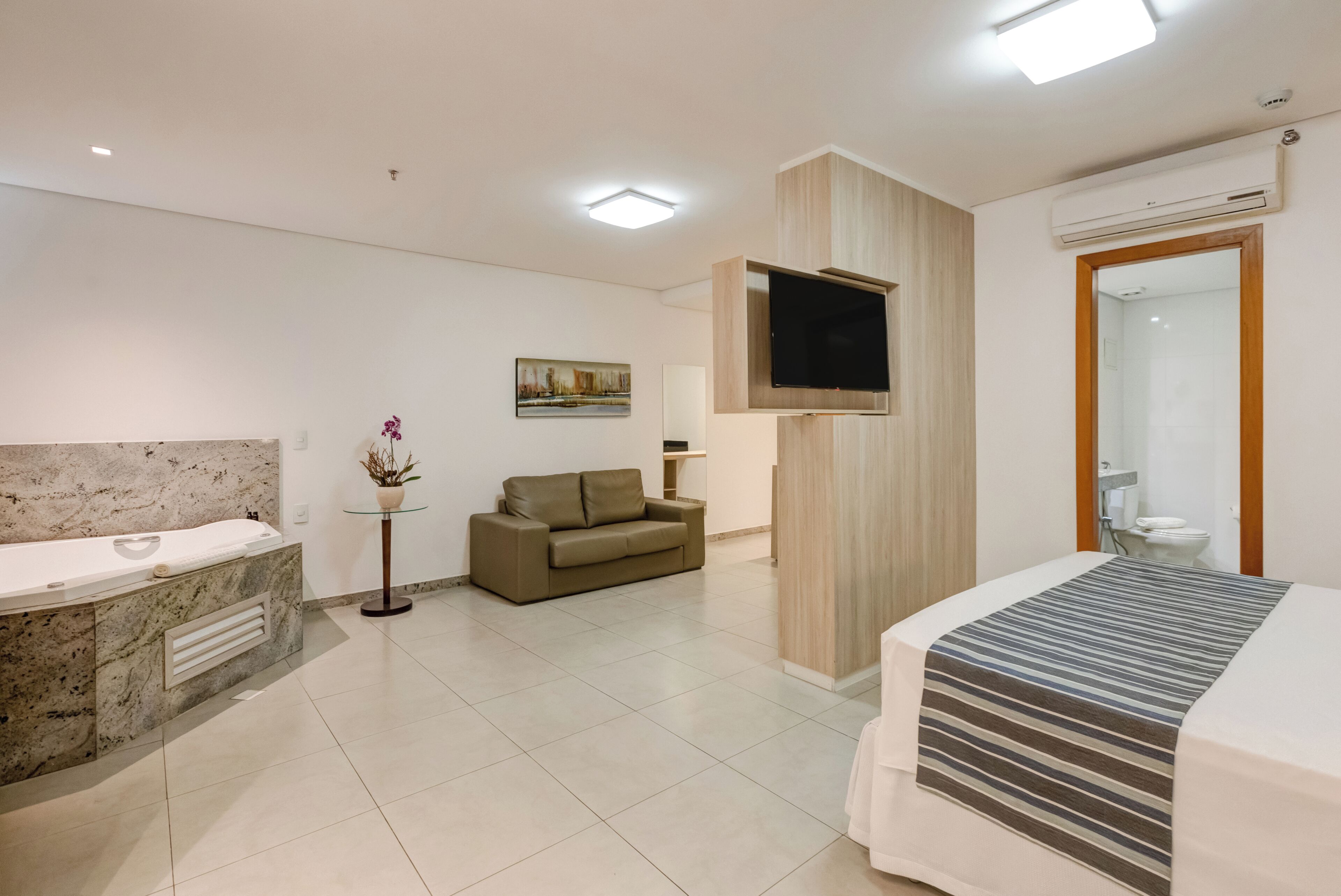 A modern hotel room with a marble bathroom, a sofa, a flat-screen TV, and a bed with a striped bedspread.