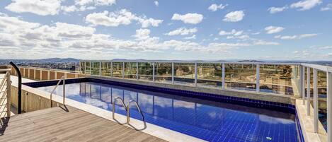 Una piscina en la azotea con cubierta de madera y barandillas de vidrio, que ofrece vistas a la ciudad y a las montañas.