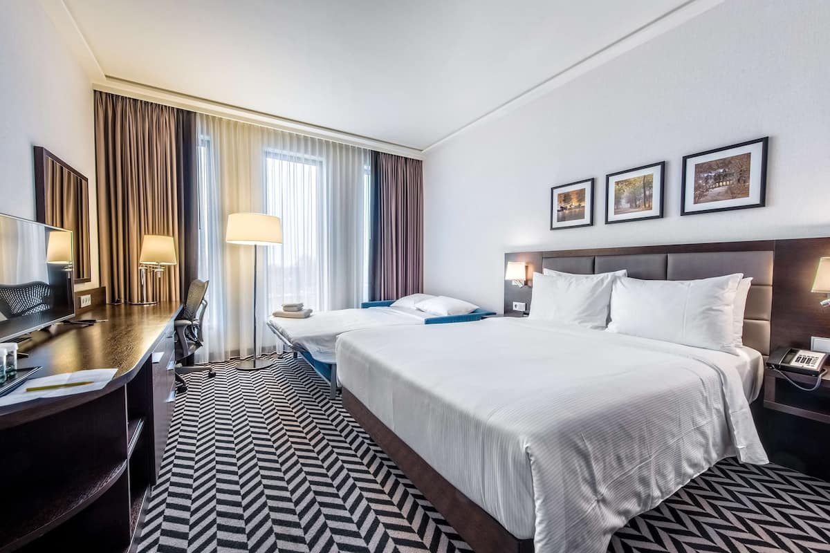 Une chambre d’hôtel avec un grand lit, un bureau, une chaise, une lampe de bureau, un téléphone et une fenêtre avec des rideaux.
